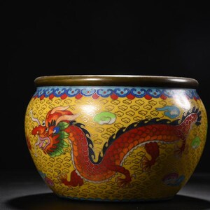 May include: A round cloisonné bowl featuring a vibrant yellow background with a repeating geometric pattern. A large red dragon with green and blue accents wraps around the exterior. The bowl has a smooth, dark metallic rim and a flat base.