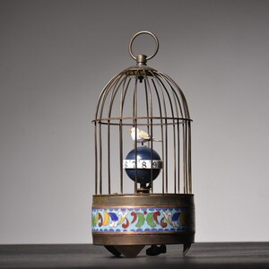 May include: A vintage birdcage clock with a white bird inside. The clock face is a blue sphere with black numbers. The base of the cage is decorated with a colorful enamel design.