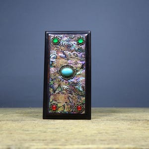 May include: A rectangular dark wood box featuring an iridescent abalone shell inlay on the front panel. The center is adorned with an oval turquoise cabochon in a filigree setting, flanked by two green and two red circular stone accents.