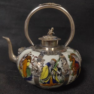 Könnte beinhalten: Eine weiße Porzellan-Teekanne mit einem silbernen Henkel und Deckel. Die Teekanne ist mit bunten bemalten Figuren und silbernen Akzenten verziert.