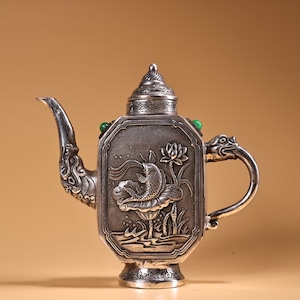 May include: Silver teapot with a dragon handle and a lid with a green stone finial. The teapot is decorated with an embossed design of a fish swimming in a lotus pond.