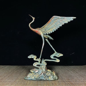 May include: A bronze sculpture of a crane with outstretched wings standing on a rock with a stylized design. The crane is painted in shades of green, red, and gold.