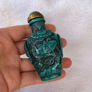 May include: A small, teal-colored, carved jade snuff bottle with a gold-colored metal lid. The bottle is decorated with intricate floral and leaf designs.