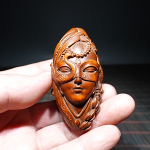 May include: A hand-carved wooden sculpture of a woman's face with a peacock feather and a snake design. The sculpture is made of dark brown wood and has a detailed design.