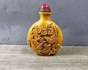 May include: A yellow snuff bottle with a crackled texture featuring relief carvings of birds and flowering branches. The bottle has a narrow neck and a rounded body, topped with a metallic cap featuring a smooth, bright red spherical finial.