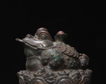 May include: A dark bronze-toned three-legged toad figurine featuring a coin in its mouth, a round object on its back, and textured skin with coin motifs. The toad rests on a circular base decorated with relief carvings and Chinese characters.