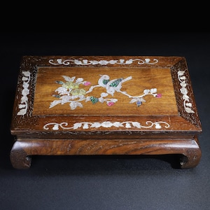 Puede incluir: Una caja rectangular de madera con acabado marrón oscuro. La parte superior presenta un diseño incrustado de un pájaro posado en una rama floreciente, con detalles de nácar. La caja se apoya en cuatro patas curvas.