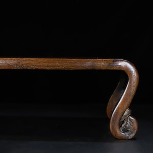 May include: A wooden stand with a dark brown finish. The stand features a curved leg with a decorative, carved floral design at the base. The top of the stand is a straight, rectangular shape.