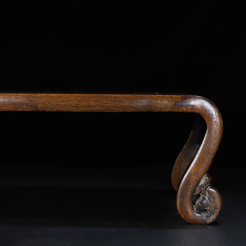 May include: A wooden stand with a dark brown finish. The stand features a curved leg with a decorative, carved floral design at the base. The top of the stand is a straight, rectangular shape.