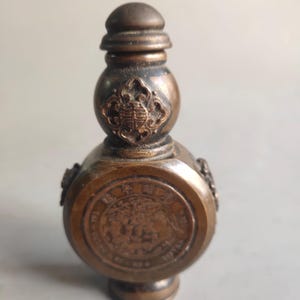 Puede incluir: Una botella de color bronce, de estilo antiguo, con cuerpo redondeado y tapón decorativo. La botella presenta diseños en relieve, incluyendo un emblema circular central y detalles laterales ornamentados. El diseño general sugiere un artículo vintage o de colección.