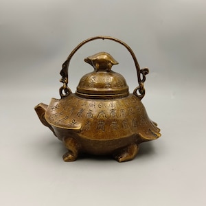 Puede incluir: Una tetera pequeña de bronce con forma de tortuga, con tapa y asa. La tetera está decorada con caracteres chinos.