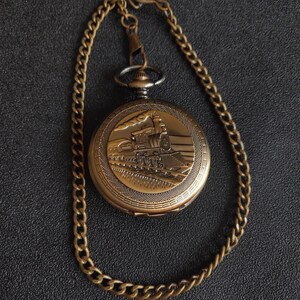 May include: A gold pocket watch with a chain. The watch face features an engraved image of a steam locomotive.