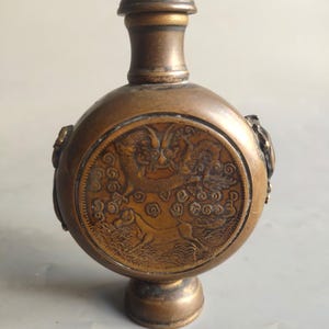 Puede incluir: Una pequeña botella redonda de latón con un diseño de dragón y nubes. La botella tiene un tapón y una pequeña base.