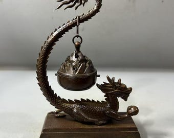 Op de afbeelding: Een bronzen wierookbrander in de vorm van een draak met een bel die aan zijn staart hangt. De draak is om een basis gewikkeld en heeft een gedetailleerd ontwerp.