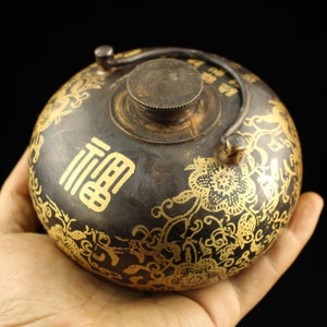 Könnte beinhalten: Eine kleine, runde, schwarze und goldene Teekanne mit Deckel und Henkel. Die Teekanne ist mit floralen Mustern und chinesischen Schriftzeichen verziert.