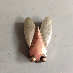 Könnte beinhalten: Ein Glücksbringer in Form einer Zikade aus Silber und Kupfer mit filigranen Details und chinesischen Schriftzeichen. Der Glücksbringer hat einen Kupferkörper und silberne Flügel.