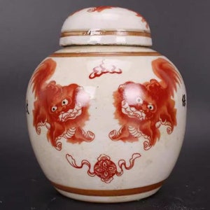 Op de afbeelding: Een witte porseleinen pot met een deksel, versierd met rood en zwart geschilderde mythische wezens. De pot heeft een gouden rand rond de boven- en onderkant.