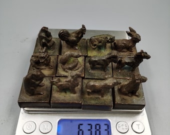 May include: A set of 12 small bronze animal figurines, each on a square base, arranged in a grid pattern on a digital scale that reads 630.3. The scale has buttons labelled "Open", "Unit", "PCS", and "Tare".