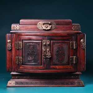 May include: A dark wood jewelry box with intricate carvings and brass accents. The box has two doors with carved figures and a brass latch. The box is sitting on a teal surface.