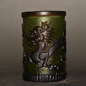 May include: A cylindrical, green glass container with a detailed, dark brown dragon carving. The container has a textured surface and a dark brown rim and base. The dragon is intricately carved, adding a touch of artistry to the piece.