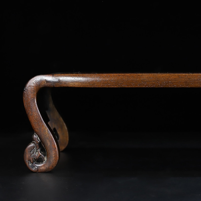May include: A dark brown wooden stand with a curved leg design. The stand has a rectangular top and a decorative, carved scroll design on the legs. The background is black.