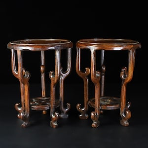 Könnte beinhalten: Zwei dunkelbraune Pflanzenständer aus Holz mit aufwendigen Schnitzereien. Jeder Ständer hat eine runde Oberseite und drei Beine.