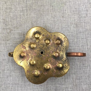 Puede incluir: Una tetera de metal de color dorado con un cuerpo en forma de flor y un asa de cobre. La tetera tiene un pequeño pico y está decorada con un diseño de peces y plantas. La tetera tiene una serie de pequeños bultos alrededor del borde.