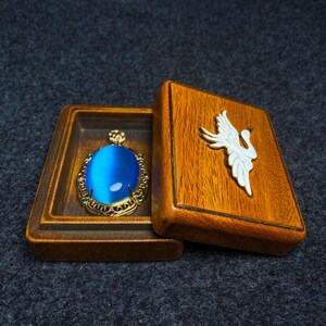 Op de afbeelding: Een blauwe edelsteen hanger met gouden accenten en rode en groene stenen in een houten doos met een wit kraanvogel ontwerp op de deksel.