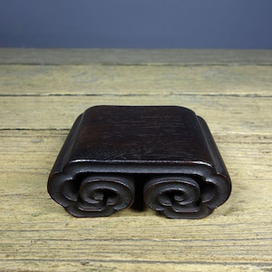 May include: A dark brown wooden stand with a scroll design. The stand is made of wood and has a smooth finish.