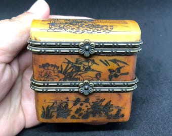 Puede incluir: Una caja rectangular de color ámbar con dos compartimentos apilados, con ilustraciones oscuras grabadas de aves, follaje y figuras. La caja está asegurada por dos bisagras de metal bronce con cierres en forma de flor en la parte frontal.