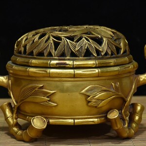 Könnte beinhalten: Ein goldener Räuchergefäß mit einem Bambus-Design. Der Deckel hat aufwendige Blattausschnitte, und der Körper ist mit Blatt- und Bambusstiel-Details verziert. Der Brenner hat drei bambusförmige Beine und zwei Griffe, was eine elegante, traditionelle Ästhetik schafft.