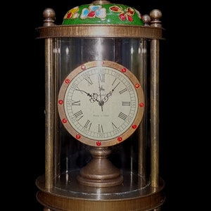May include: A brass clock with a glass dome and a Roman numeral clock face. The clock face has red accents and the text "Made in 1886". The clock has a green top with colorful floral designs.