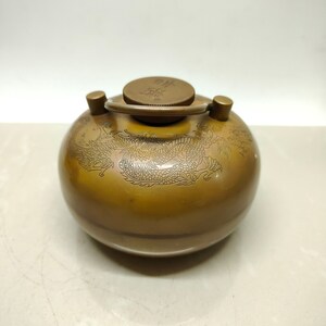 May include: A small, round, brass container with a lid and a handle. The container is decorated with an engraved design of a dragon. The lid has Chinese characters on it.