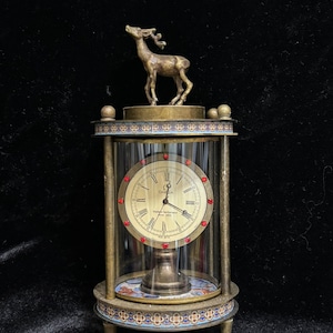 May include: A vintage-style, gold-toned table clock with a glass dome and a deer figurine on top. The clock face is white with black Roman numerals and the words "Omega Made in Switzerland since 1848".