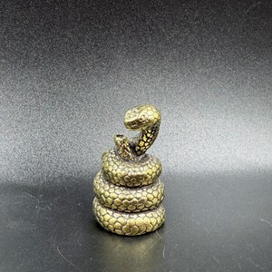 May include: A gold-coloured metal figurine of a coiled snake with its head raised. The snake is detailed with scales and has a realistic appearance.