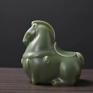May include: A green ceramic horse figurine with a smooth, glossy finish. The horse is in a seated position, with a detailed mane and saddle. The figurine is a solid, decorative piece.