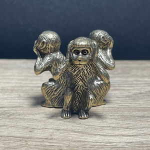 Puede incluir: Tres pequeños monos dorados, cada uno cubriendo un sentido diferente: uno se tapa los ojos, otro se tapa las orejas y el último se tapa la boca. Los monos están dispuestos en círculo, mirando hacia afuera.