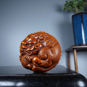 Op de afbeelding: Een handgesneden houten sculptuur van een draak, met ingewikkelde details van schubben en een woeste uitdrukking. Het beeld is bruin en heeft een ronde vorm.
