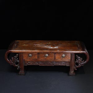 May include: A dark brown wooden table with three drawers and ornate carvings. The table has curved sides and decorative dragon-like figures on the legs. The top surface features inlaid floral designs. The drawer pulls are circular and metallic.