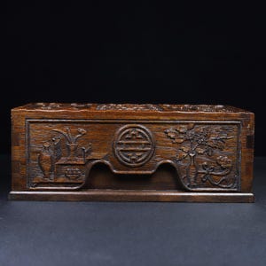 May include: A rectangular wooden box with intricate carvings. The box features floral and vase designs on the front, with a central circular emblem. The wood has a rich, dark brown color, and the carvings add depth and texture.