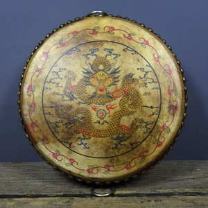 May include: A round, leather-covered drum with a brass rim and a painted design of a dragon in red, yellow, and black. The dragon is surrounded by clouds and a floral border.