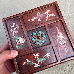 May include: A square wooden box featuring dark brown wood grain and inlaid floral motifs made of mother-of-pearl and colored stone. The center displays a circular metal ornament with a red stone surrounded by small turquoise-colored beads.