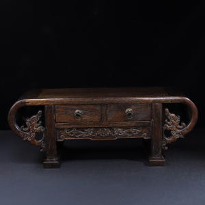 May include: A dark brown wooden miniature cabinet with two drawers and ornate dragon carvings on the sides. The cabinet has a curved top and decorative pulls on the drawers. The legs are square and sturdy.