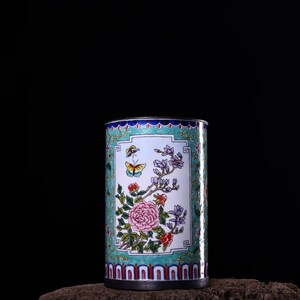 Op de afbeelding: Een cilindrische blauwe en witte emaille vaas met een bloemenontwerp met roze bloemen, groene bladeren en een vlinder. De vaas heeft een gouden rand en een decoratieve band van rood, blauw en wit rond de onderkant.