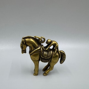 Puede incluir: Una figurita de metal dorado de un caballo con un mono montado en su lomo. El caballo tiene una silla de montar y una brida, y el mono se agarra a la crin del caballo.