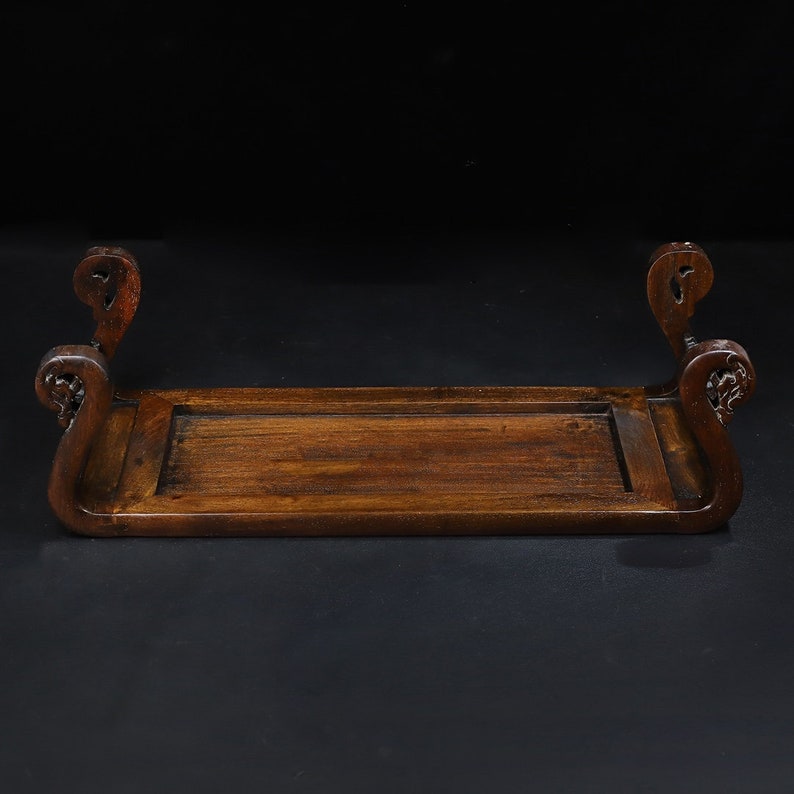 May include: A dark brown wooden display stand with a rectangular base and curved, ornate ends. The stand has a carved design on the ends and is set against a black background. The stand is likely used for displaying decorative items.