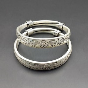 May include: Two silver bangles with a floral design. The bangles are stacked on top of each other.