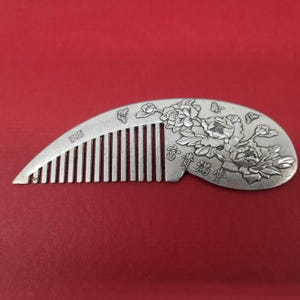 Puede incluir: Un peine de plata con un diseño adornado de flores y mariposas. El peine tiene una fila de dientes y está decorado con caracteres chinos.