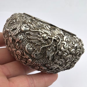 Puede incluir: Un brazalete ovalado plateado con intrincados diseños de dragones y nubes. Los grabados detallados cubren toda la superficie, reflejando un estilo asiático tradicional. El metal presenta un aspecto ligeramente envejecido.