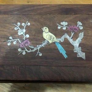 Può includere: Scatola rettangolare in legno scuro. Il coperchio presenta un intarsio di un uccello giallo posato su un ramo con fiori rosa e piume blu. Accenti decorativi agli angoli.